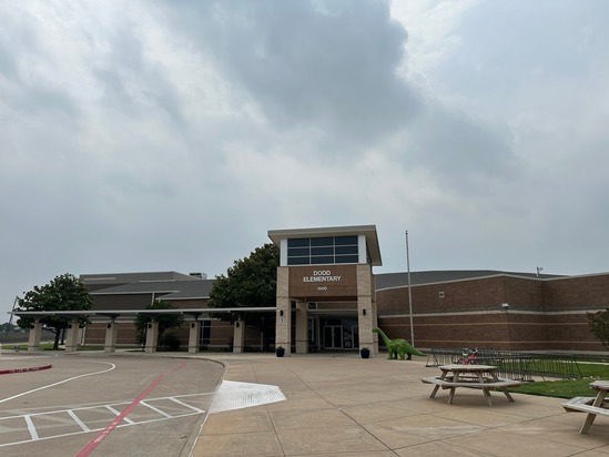 Front exterior view of Dodd Elementary School with a cloudy sky overhead, showing the main entrance, parking area, sidewalk, picnic tables, and a green dinosaur statue near the building.