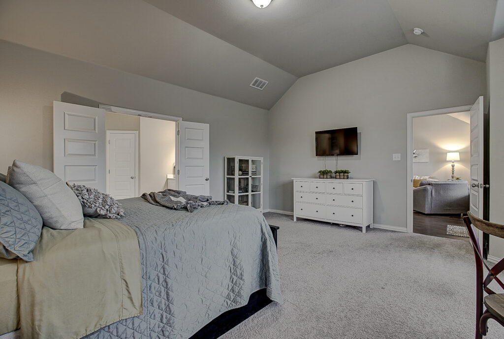 View from bed across the primary bedroom showing double doors to bathroom and TV