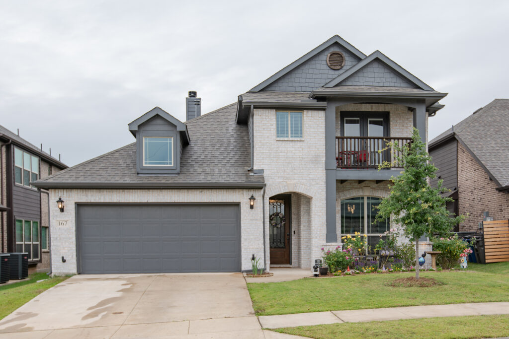 Front exterior of a two-story Bloomfield home in Grand Heritage East featuring white brick, covered balcony, landscaped yard, and cul-de-sac location in Lavon TX