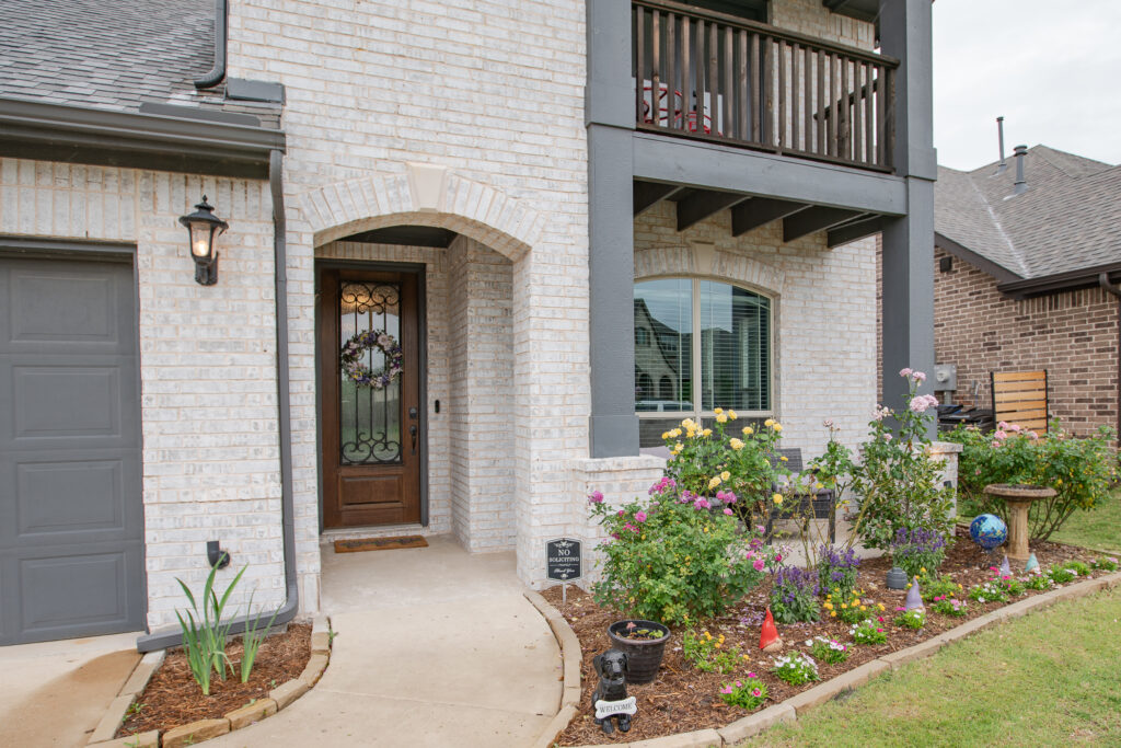 Covered front porch entry with custom wood and iron door, flower beds, and balcony detail at a Lavon TX home